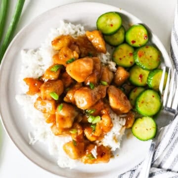 Overhead view of honey garlic chicken served on a bed of rice, on a white plate, with a side of cucumber salad.