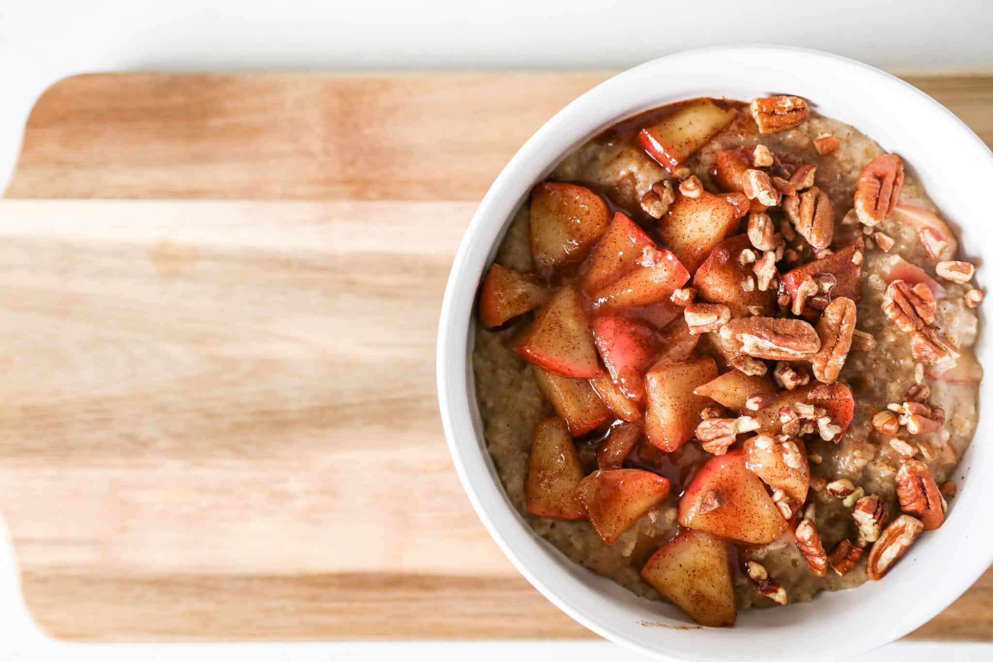Apple Cinnamon Oatmeal topped with chopped pecans in white bowl sitting on a cutting board