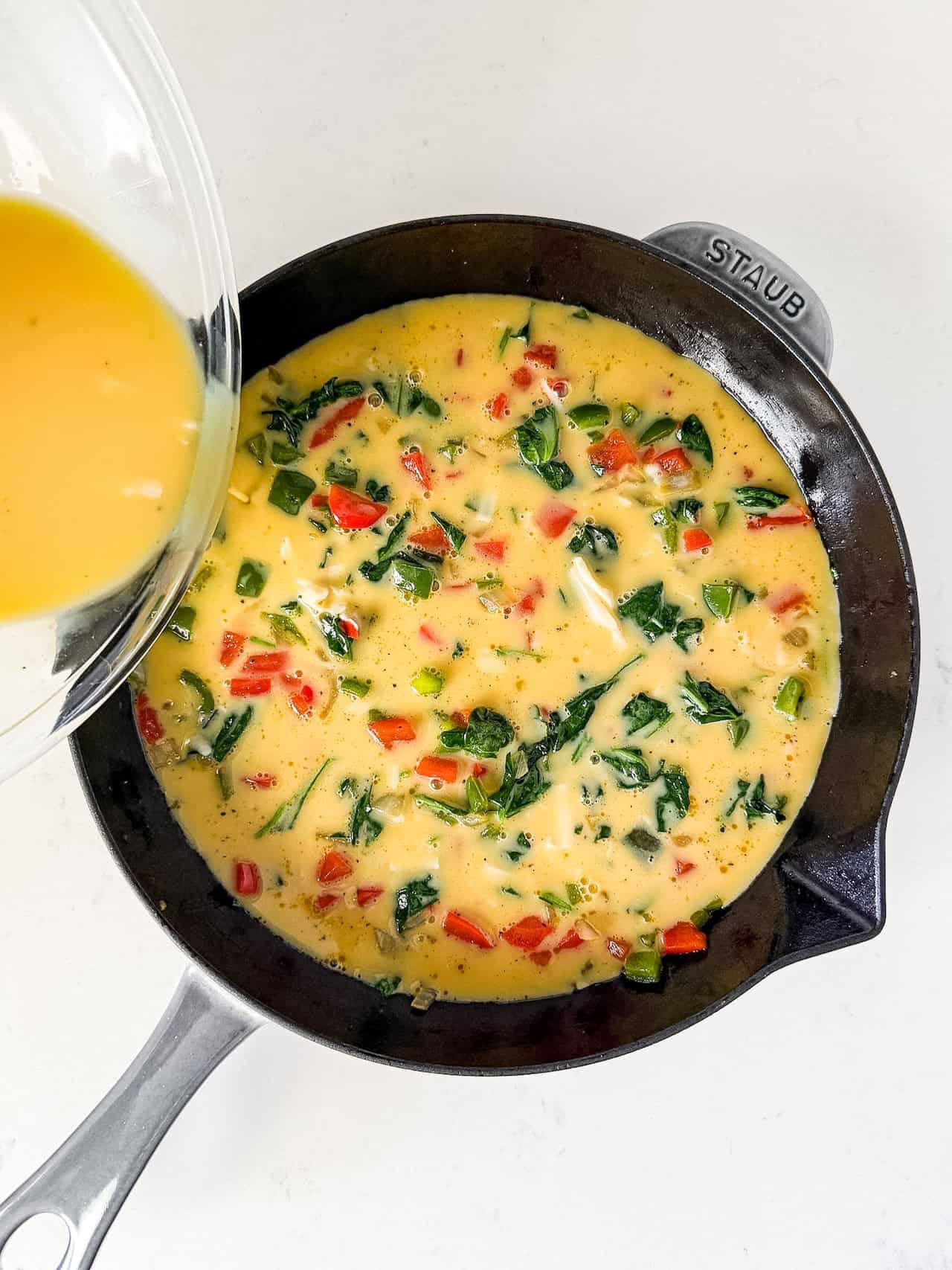Eggs being poured over vegetables in a skillet.