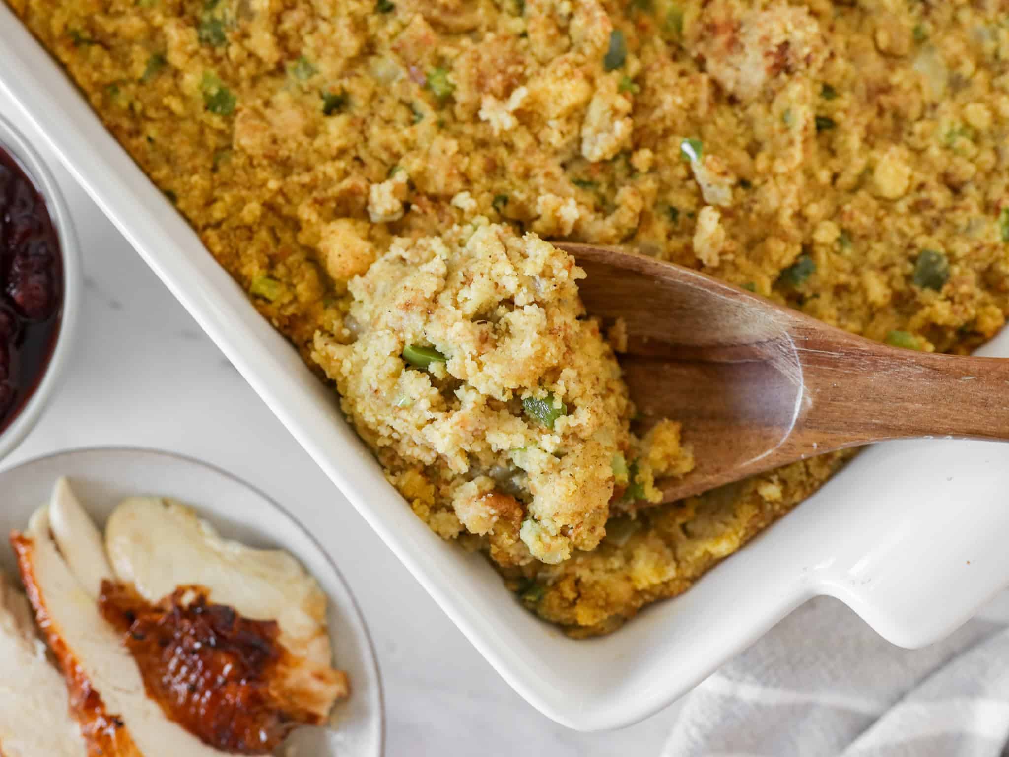 Overhead view of cornbread dressing in a white casserole dish with a wooden spoon scooping some out.