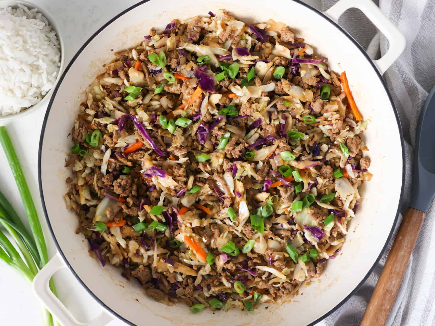 Overhead view of egg roll in a bowl in a large skillet with green onions, rice, and a wooden spoon on the side.