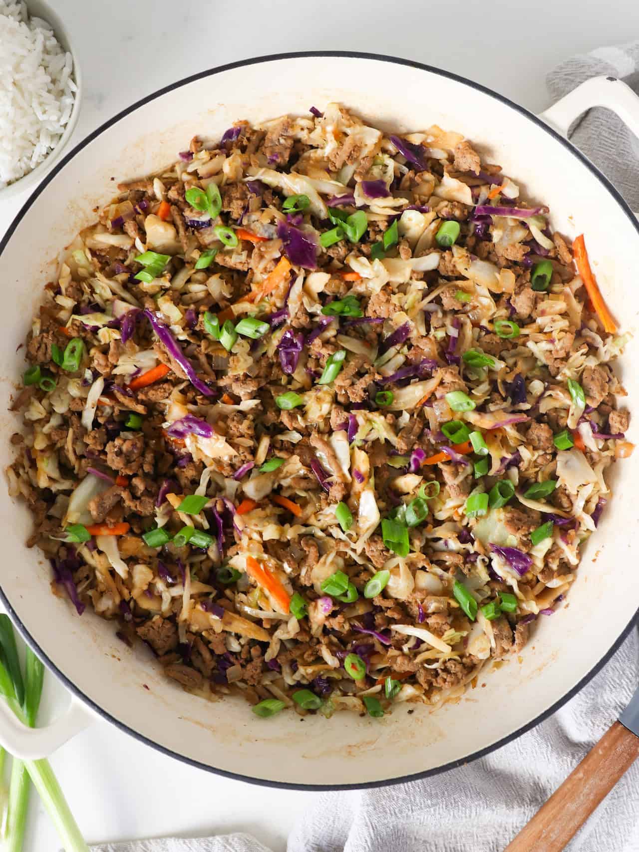 Overhead view of egg roll in a bowl in a large skillet with green onions, rice, and a wooden spoon on the side.