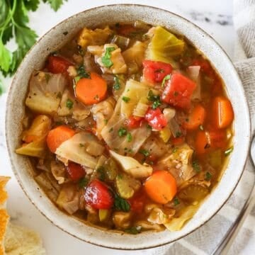 Overhead view of cabbage soup in a serving bowl with a spoon, parsley, and bread on the side.