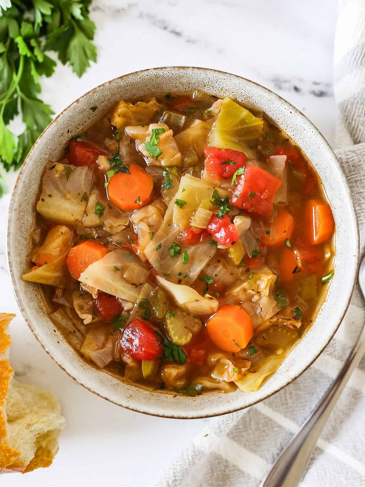 Overhead view of cabbage soup in a serving bowl with a spoon, parsley, and bread on the side.