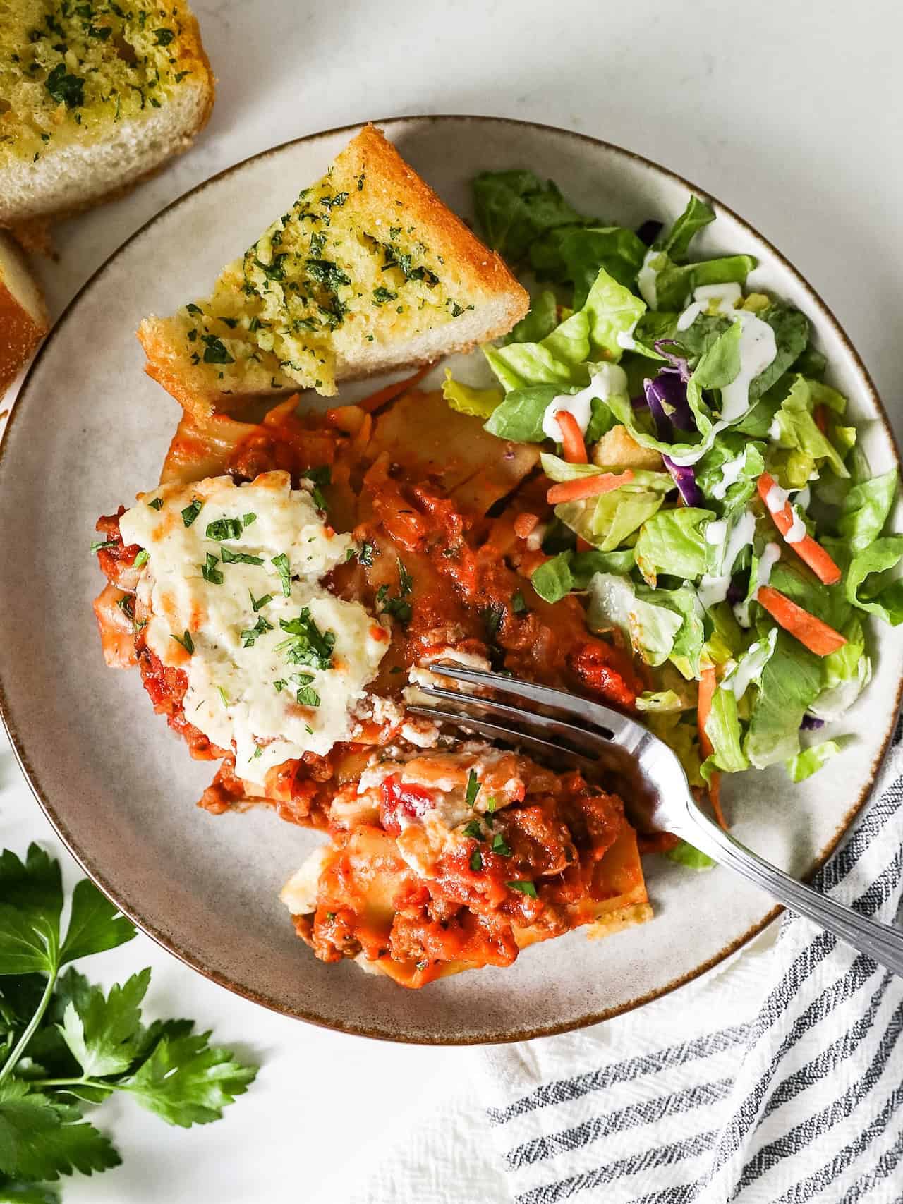 A white plate with a serving of skillet lasagna, a side salad, and a piece of garlic bread.
