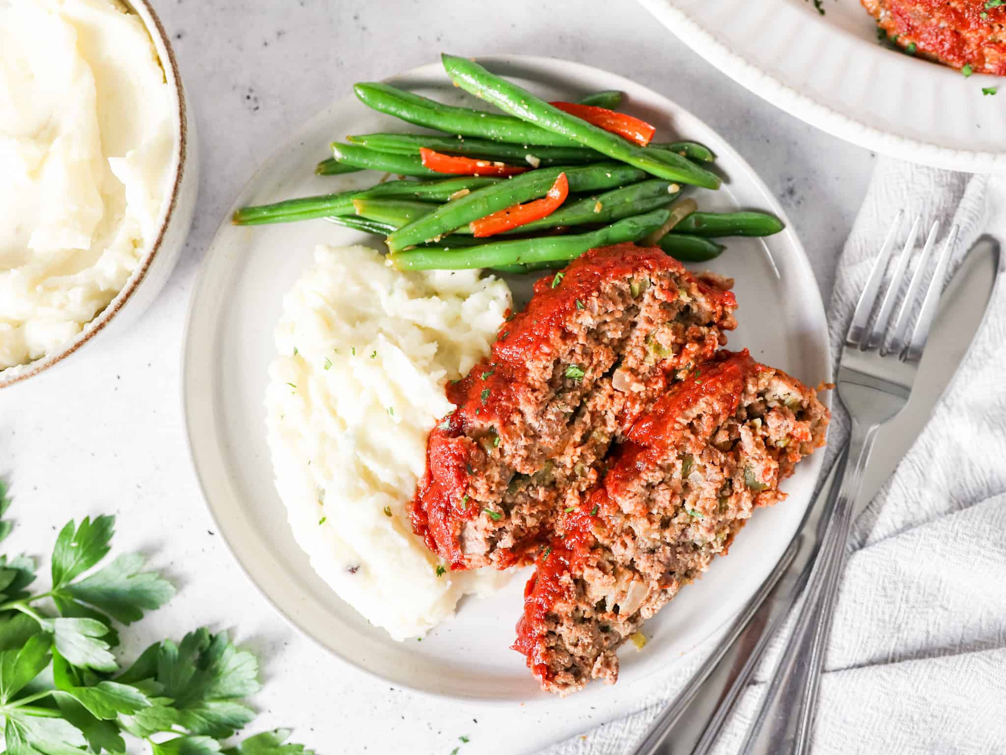 Two slices of meatloaf on a serving plate with mashed potatoes and green beans on the side.