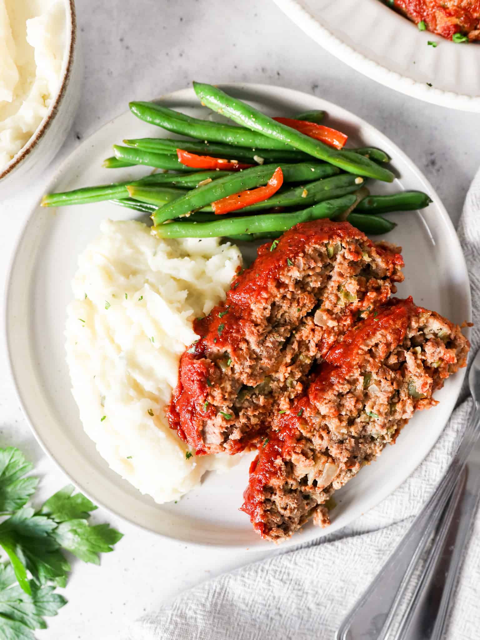 Two slices of meatloaf on a serving plate with mashed potatoes and green beans on the side.