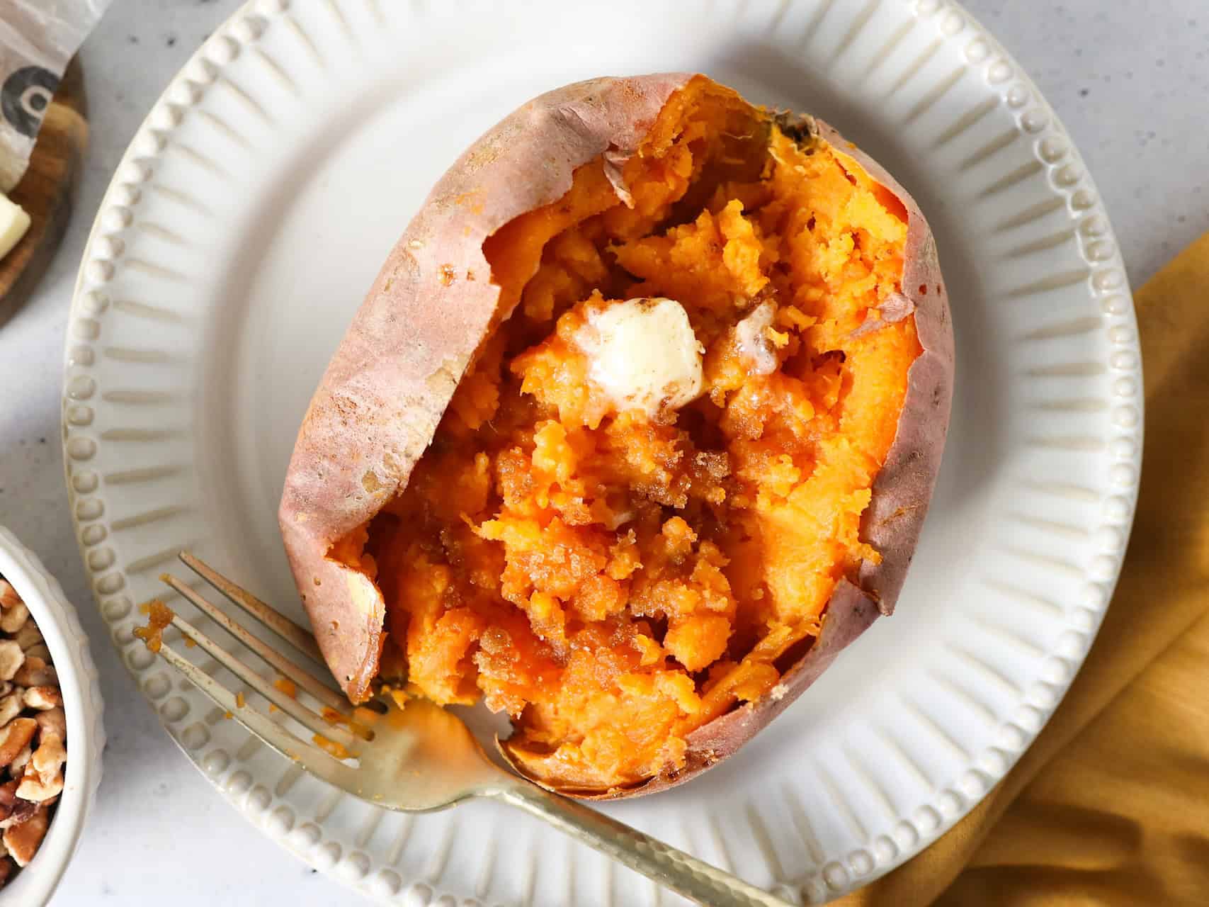Overhead view of one baked sweet potato on a white plate with a fork and napkin on the side.
